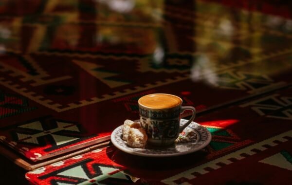 A traditional Turkish coffee served in an ornate cup with a piece of sweet dessert on a decorative plate, set against a vibrant patterned tablecloth