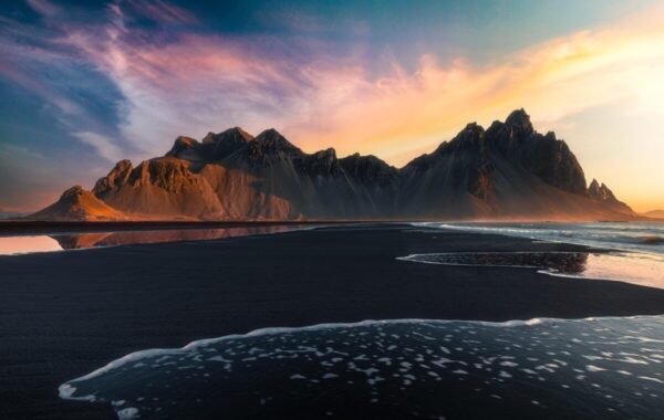 Stunning beach landscape featuring dramatic mountain silhouettes at sunset, with gentle waves lapping against the dark sand shoreline and colorful skies reflecting in the water