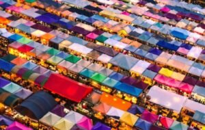 Aerial view of a vibrant outdoor market filled with colorful tented stalls, showcasing a lively atmosphere and a variety of goods illuminated by evening lights