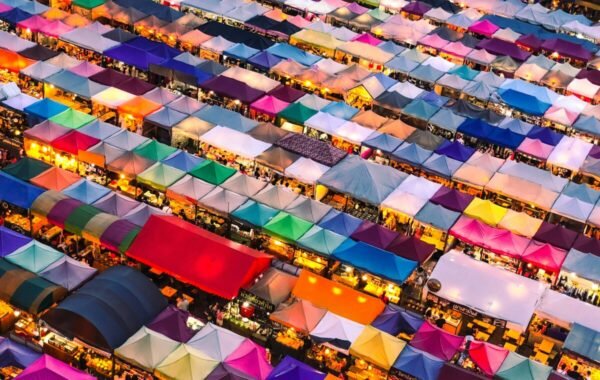 Aerial view of a vibrant outdoor market filled with colorful tented stalls, showcasing a lively atmosphere and a variety of goods illuminated by evening lights