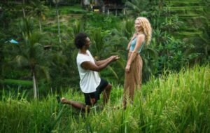 A joyful couple in a lush green field, with a man kneeling in a proposal while holding a ring, and a woman smiling in anticipation
