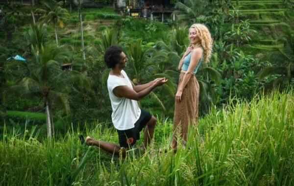A joyful couple in a lush green field, with a man kneeling in a proposal while holding a ring, and a woman smiling in anticipation