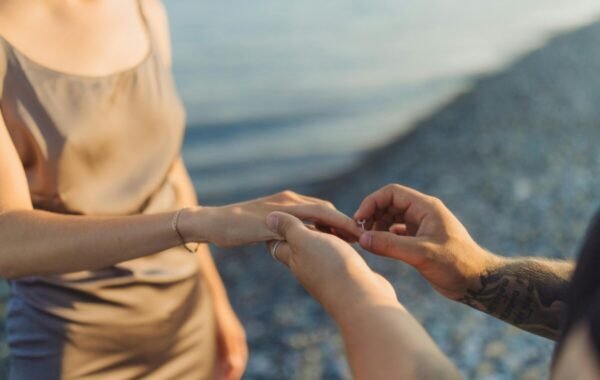 A person proposing with an engagement ring, placing it on the hand of their partner, set against a serene beach backdrop
