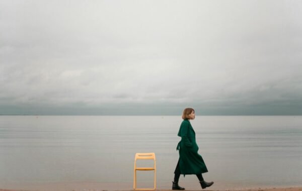 A lone woman in a green coat walks along a serene beach, with a yellow chair beside her, set against a backdrop of calm water and overcast skies, capturing the essence of solo female travel