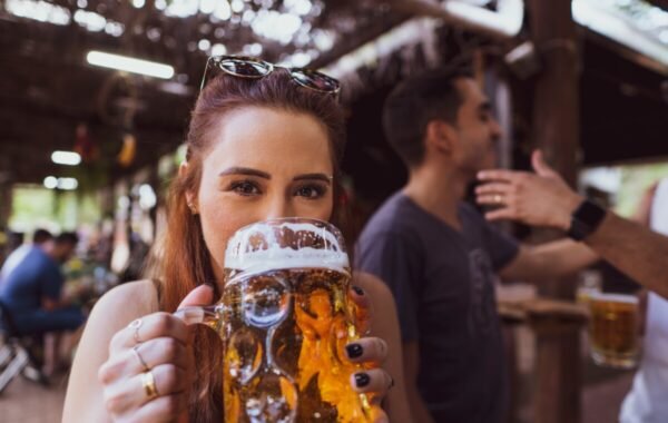 A woman smiles while holding a large stein of golden beer at a lively outdoor beer garden in Germany, with friends and tables in the background