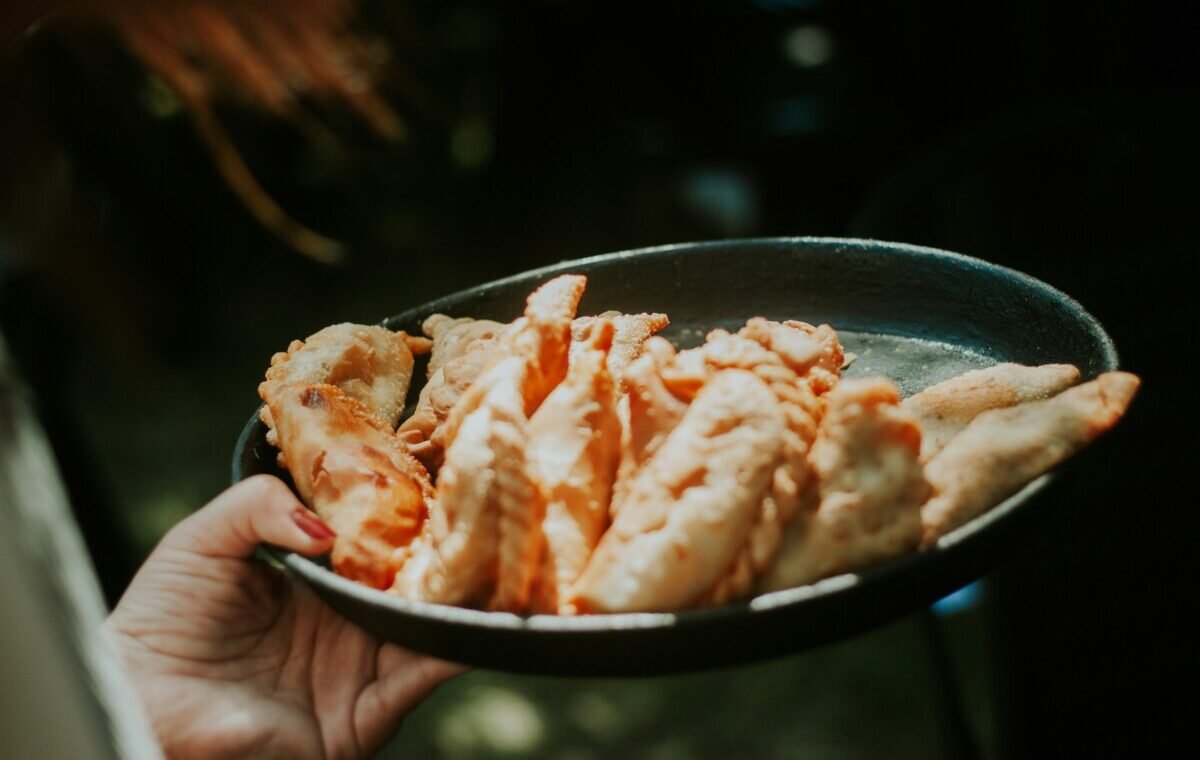 A hand holding a plate of golden-brown empanadas, showcasing a variety of shapes and textures, in a warm, natural setting