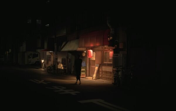 A dimly lit street in Japan at night, featuring traditional storefronts and subtle neon lights, with a lone figure walking past