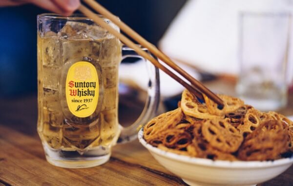 A glass of Suntory Whisky, filled with ice, sits on a table next to a bowl of crispy lotus root snacks, with chopsticks poised to pick one