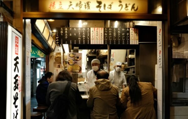 Busy street food stall in Osaka serving traditional soba noodles, featuring chefs in white uniforms and customers enjoying meals at the counter