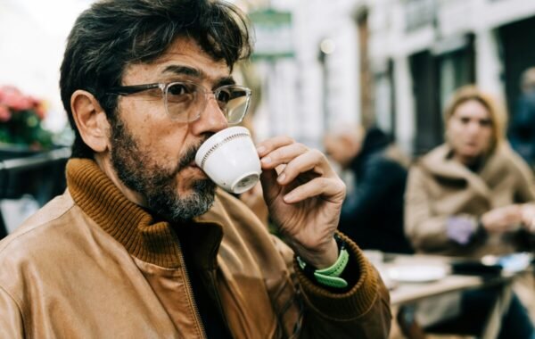 Man enjoying a traditional Italian espresso at a café in Rome, dressed in a brown leather jacket with stylish glasses, as another person relaxes in the background