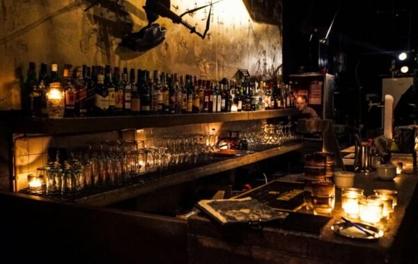 A dimly lit bar showcasing an extensive selection of whiskey bottles on the shelves, with illuminated candles and glassware on the counter, creating a cozy atmosphere for food and drink enthusiasts