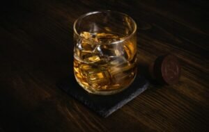 Glass of whiskey with ice cubes on a slate coaster, set against a dark wooden background, highlighting the rich amber color and inviting presentation of the drink
