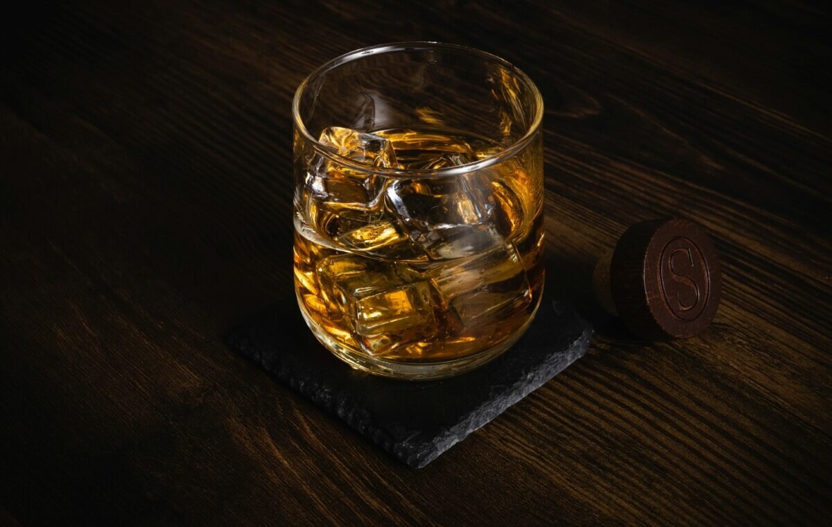 A glass of whiskey with ice cubes sits on a slate coaster, complemented by a round wooden token, against a dark wooden background