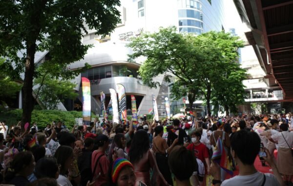 A large crowd celebrates at an outdoor event, with rainbow flags and banners visible, suggesting lgbt travel thailand
