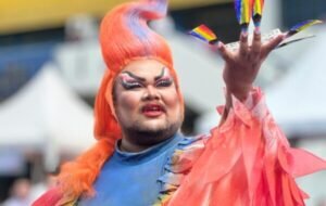 A drag performer with bright orange hair and rainbow-colored nails holds up a small rainbow flag