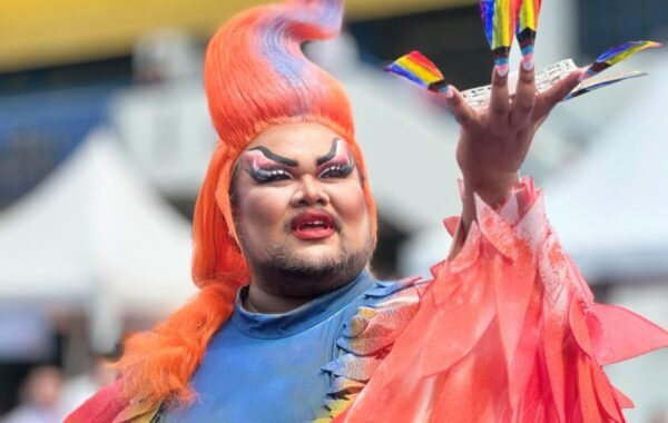 A drag performer with bright orange hair and rainbow-colored nails holds up a small rainbow flag