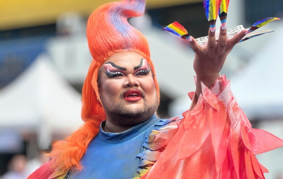A drag performer with bright orange hair and dramatic makeup holds up a hand with rainbow-painted nails, possibly representing lgbt travel thailand