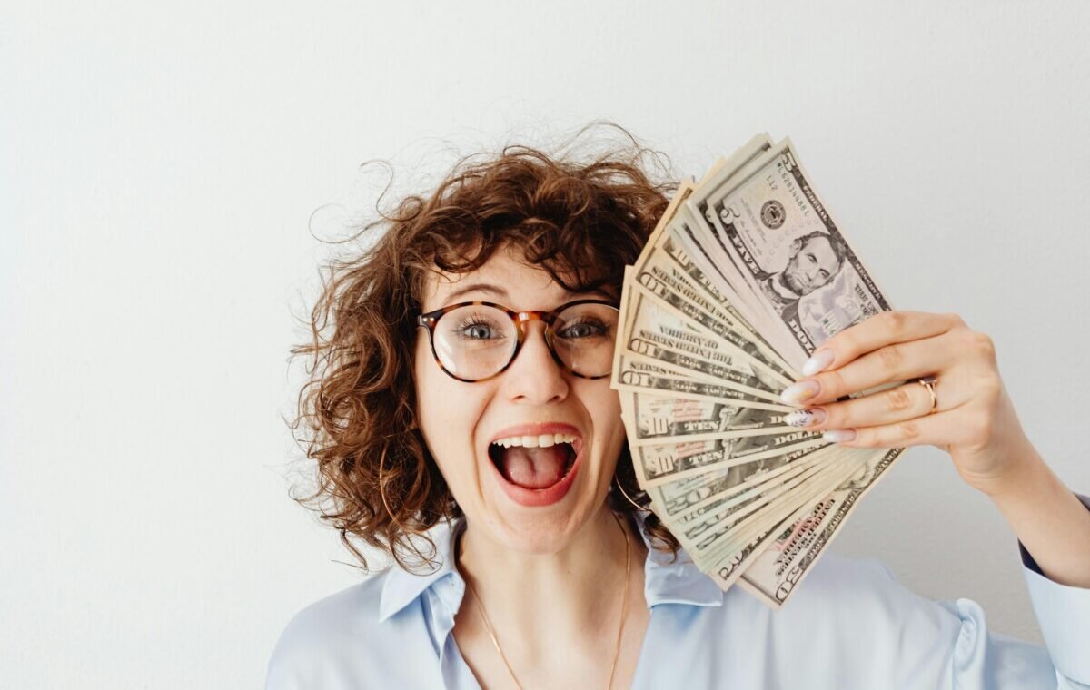 A woman with curly hair and glasses excitedly holds a fan of US dollar bills, suggesting success with side hustles traveling