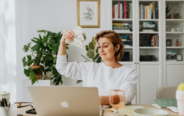 A woman holds a fan of money while working on her laptop, suggesting side hustles traveling