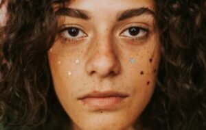 A young woman with freckles and star glitter on her face looks directly at the camera