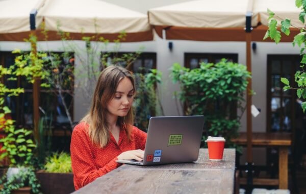 A woman in a red polka dot shirt works on her laptop at an outdoor cafe, with lush greenery in the background